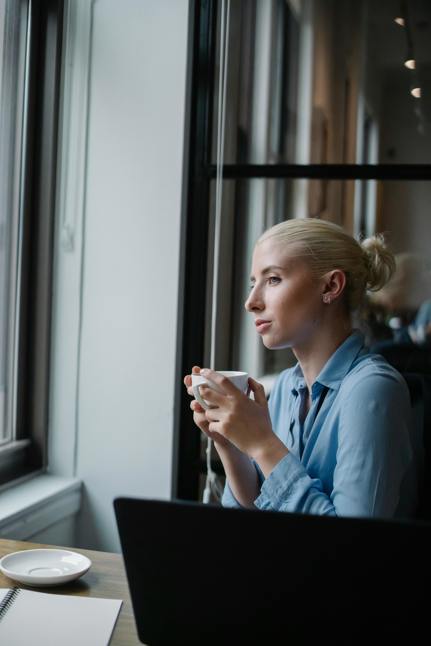 a young woman sitting in a cafe with a coffee in hand, looking outside the window. 